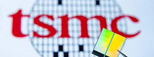 Tweezers hold a small microchip in front of a blurred TSMC logo.